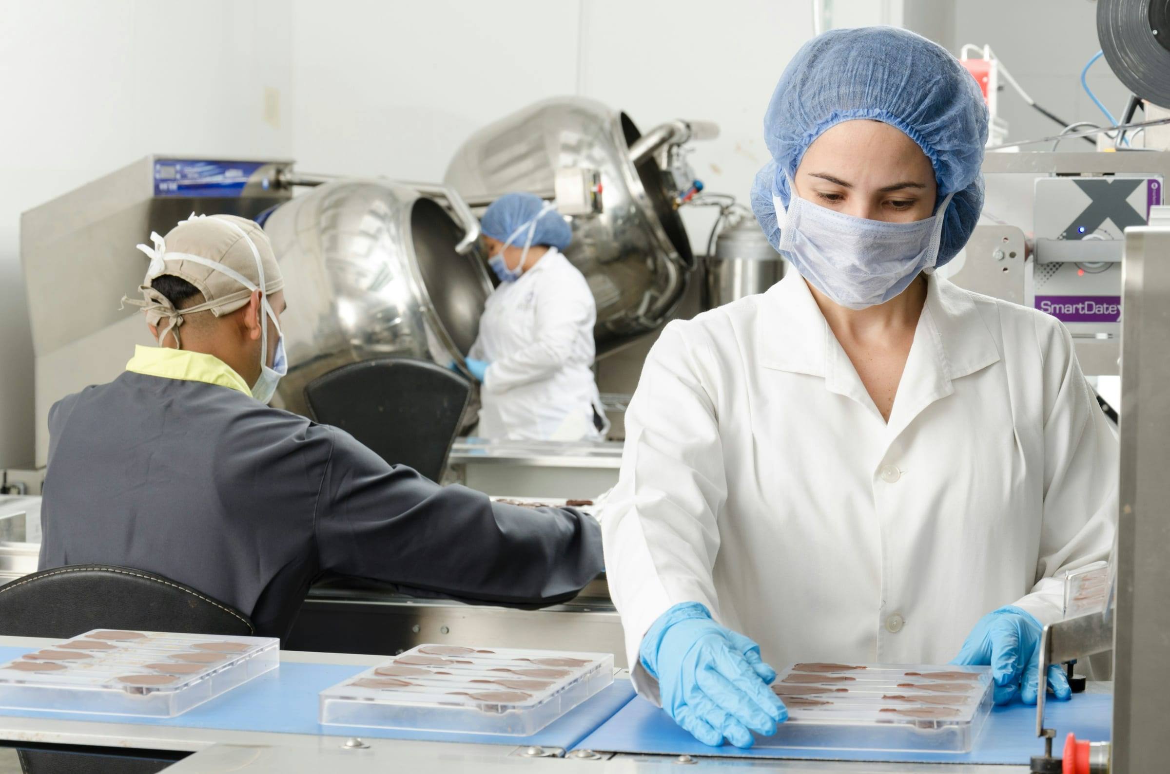 Food production worker in a blue hairnet, face mask, white coat and blue gloves handling trays of chocolate on a conveyor, with coworkers and stainless steel kettles in the background.