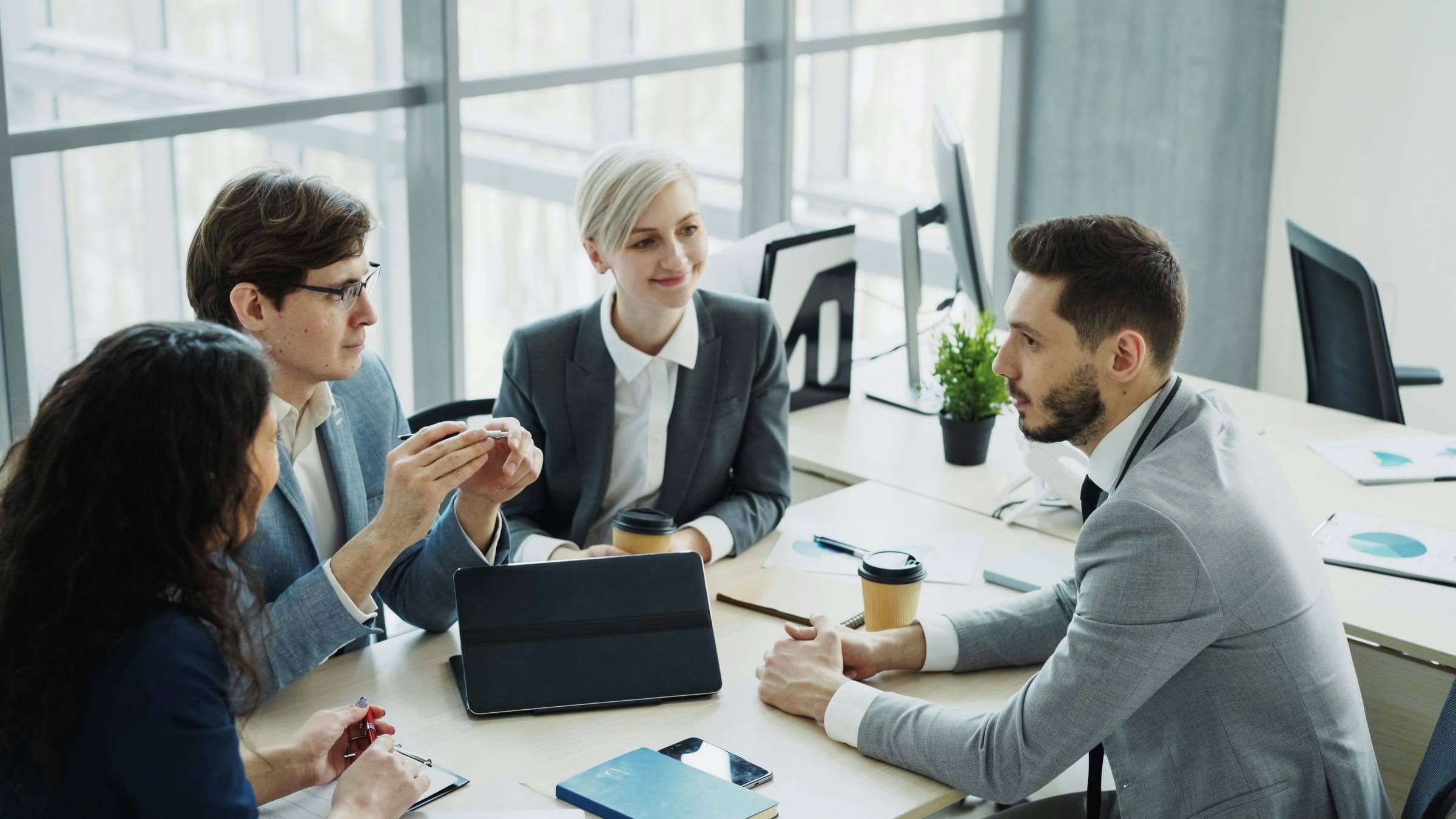 Four professionals in business attire talking around a table in a bright office, with a tablet and coffee cups.