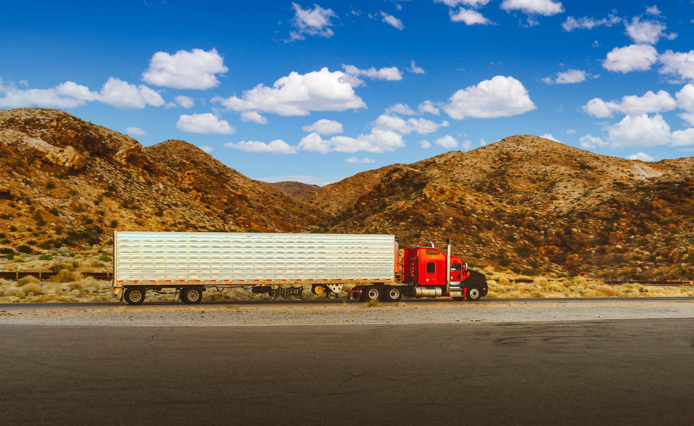 Red semi-truck pulling a long white trailer along a desert highway in Nevada, with rocky mountains and a blue sky with clouds.