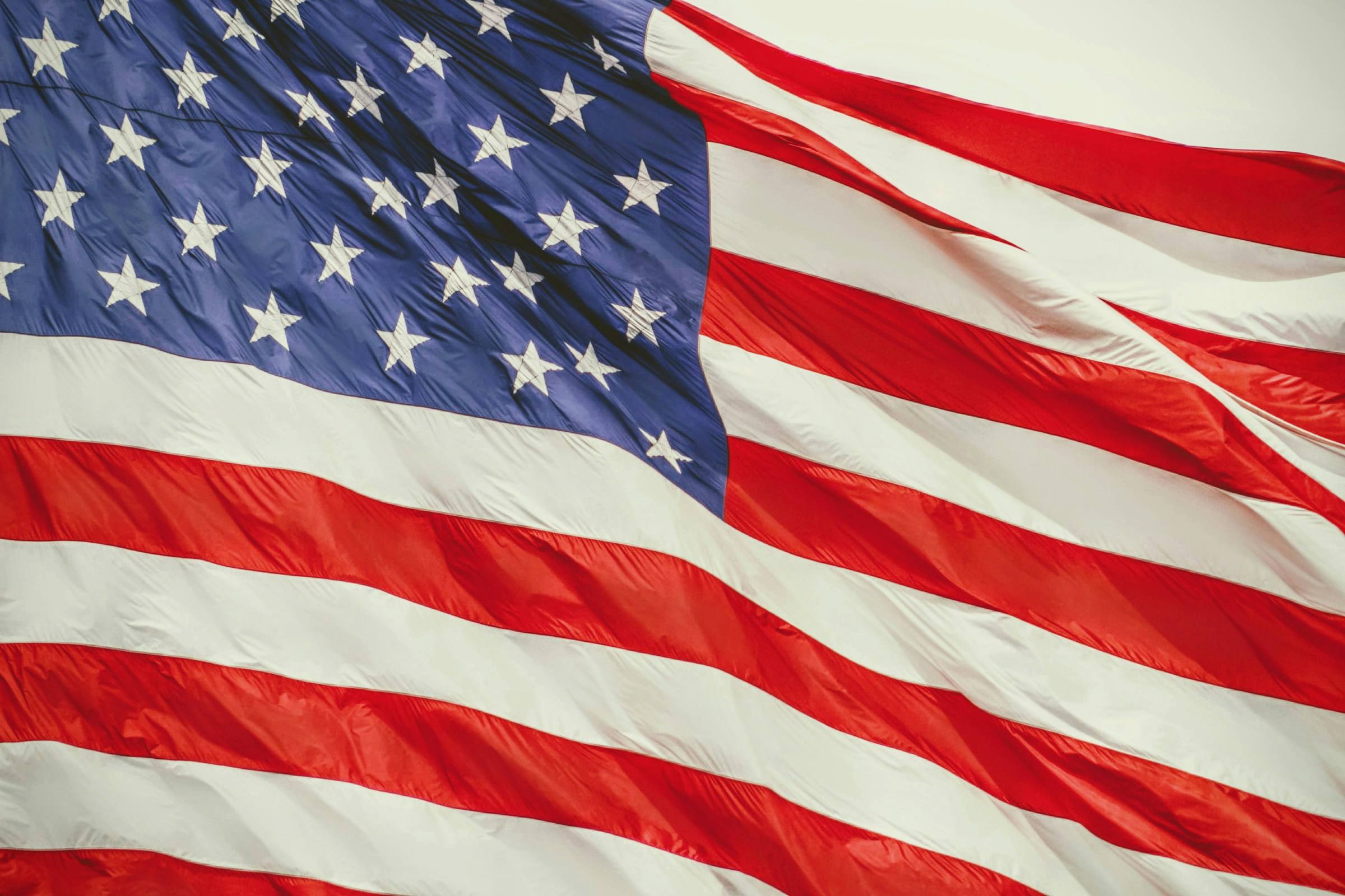 Primer plano de una gran bandera de Estados Unidos ondeando al viento, con estrellas blancas sobre un campo azul en la parte superior izquierda y franjas rojas y blancas que cruzan el encuadre en diagonal.